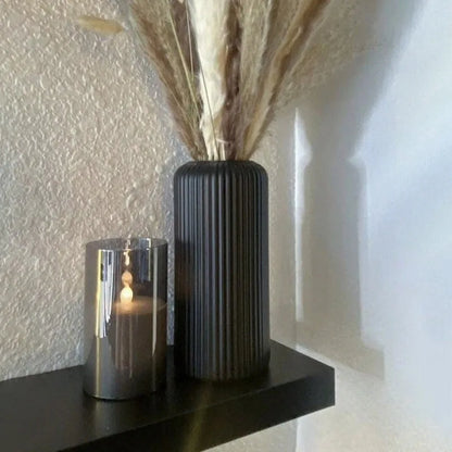 Decorative vase with dried plants and candle on a shelf against a textured wall.