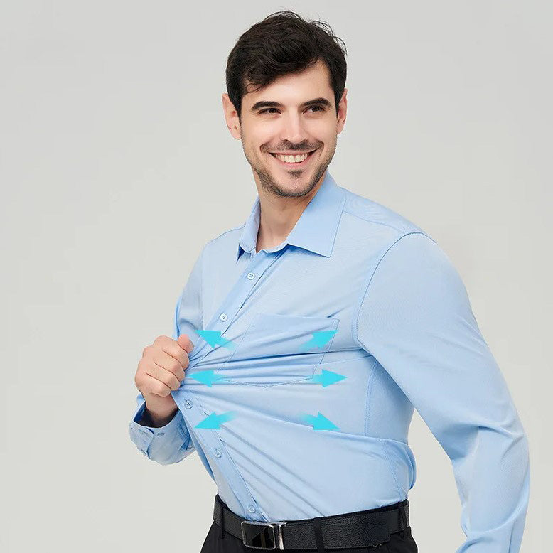 A man turned sideway standing with a smile, wearing a light blue button up dress shirt he's pulling on, dark blue pants and a black belt.