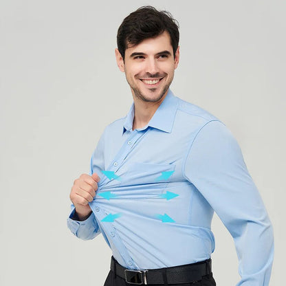 A man turned sideway standing with a smile, wearing a light blue button up dress shirt he's pulling on, dark blue pants and a black belt.